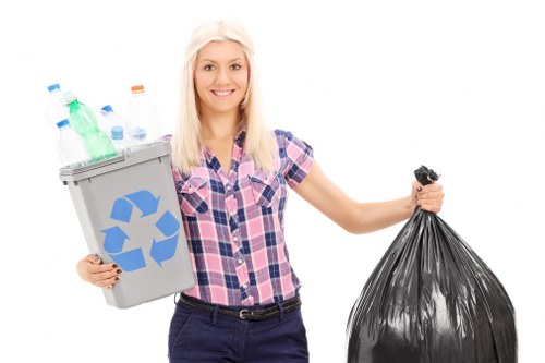 A young woman with long blonde hair smiling at the camera, wearing a short-sleeved, pink and blue plaid shirt and dark trousers, stands outdoors against a plain white background. She is holding a small grey recycling bin filled with various used plastic bottles, including transparent, green, and orange caps, in her left hand. The bin has a blue recycling symbol on the front. In her right hand, she holds a large black plastic rubbish bag, which appears to be filled with waste material. The scene emphasizes waste collection and rubbish disposal, aligned with rubbish removal services offered by companies like Office Clearance Turnham Green, and subtly references environmental management in a residential or commercial setting, possibly in Turnham Green or nearby in West London.