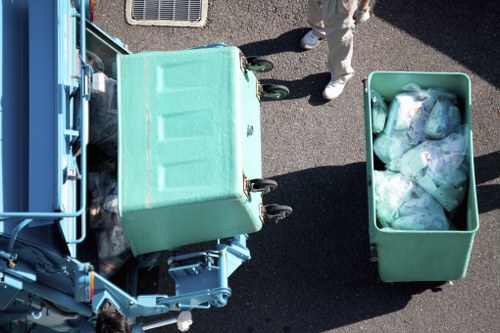 An overhead view of a light teal commercial waste bin positioned next to a larger waste collection vehicle on a paved surface, with a person standing nearby wearing light-colored trousers and shoes. The larger vehicle's open rear loading area reveals its internal compartments, and the smaller bin, filled with transparent plastic bags containing mixed waste, is attached to the vehicle via a hydraulic lifting mechanism. The scene appears to be part of a rubbish removal operation in an outdoor setting, possibly within a town or residential area near Turnham Green, with the company Office Clearance Turnham Green providing rubbish disposal services. The environment is well-lit, casting distinct shadows, and the image captures the practical aspects of waste collection and bin handling relevant to local rubbish removal services.