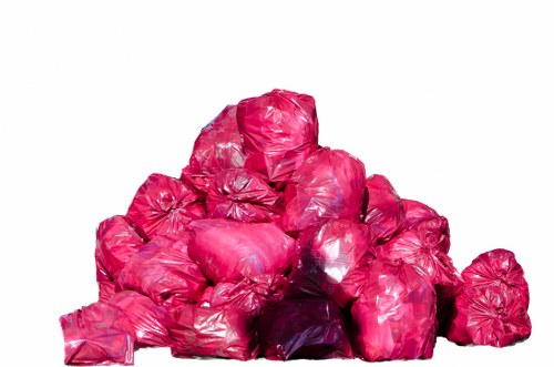 A pile of numerous bright pink plastic rubbish bags stacked outdoors against a plain white background. The bags are filled with waste, with some showing slight creases and shiny surface textures. They vary in size but are generally rounded and tightly packed, with a few bags partially leaning against each other. The plastic material appears flexible and glossy, reflecting light in certain areas. The overall arrangement suggests a collection of waste awaiting removal, typical of a rubbish clearance process in a residential or commercial setting, possibly in Turnham Green or surrounding areas. The absence of additional objects or environmental features emphasizes the focus on this heap of waste bags, which could be part of a larger rubbish removal service undertaken by Office Clearance Turnham Green.