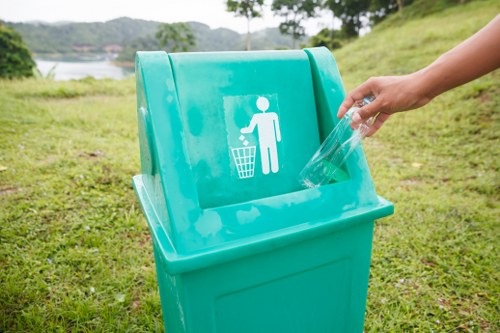 A person discards a clear plastic bottle into a large, light blue recycling bin positioned on a grassy area outdoors, with a scenic background of trees and a body of water in the distance. The bin features a white symbol of a human figure dropping waste into a bin and is situated on a paved or grassy surface. The scene appears bright with natural daylight, suggesting a park or natural setting near Turnham Green. The presence of the recycling bin reflects waste management practices related to environmental awareness, aligning with rubbish removal services provided by Office Clearance Turnham Green. The bin's smooth plastic surface has a slightly reflective finish, and the personâ€™s hand is visible, placing the bottle carefully into the designated compartment. The background includes some foliage and an open landscape, emphasizing the outdoor environment typical of local community spaces or green areas in the vicinity, subtly supporting local SEO signals for rubbish disposal services near the area.