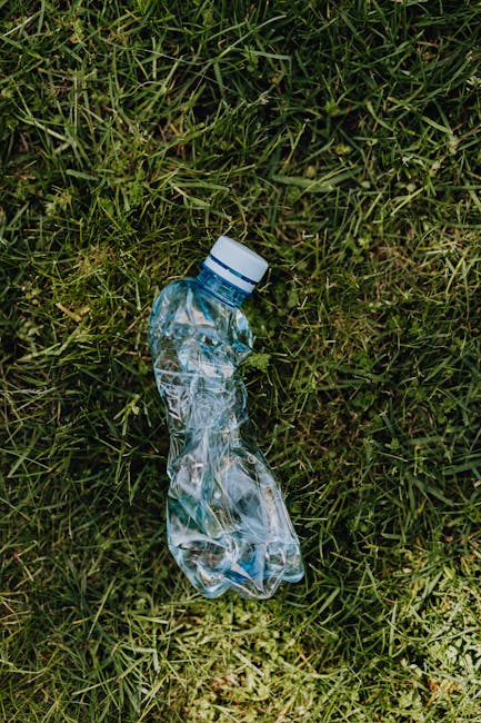 A discarded clear plastic water bottle lies on a patch of green grass with a mixture of short and slightly longer blades. The bottle appears crumpled and empty, with a white cap securely fitted on top