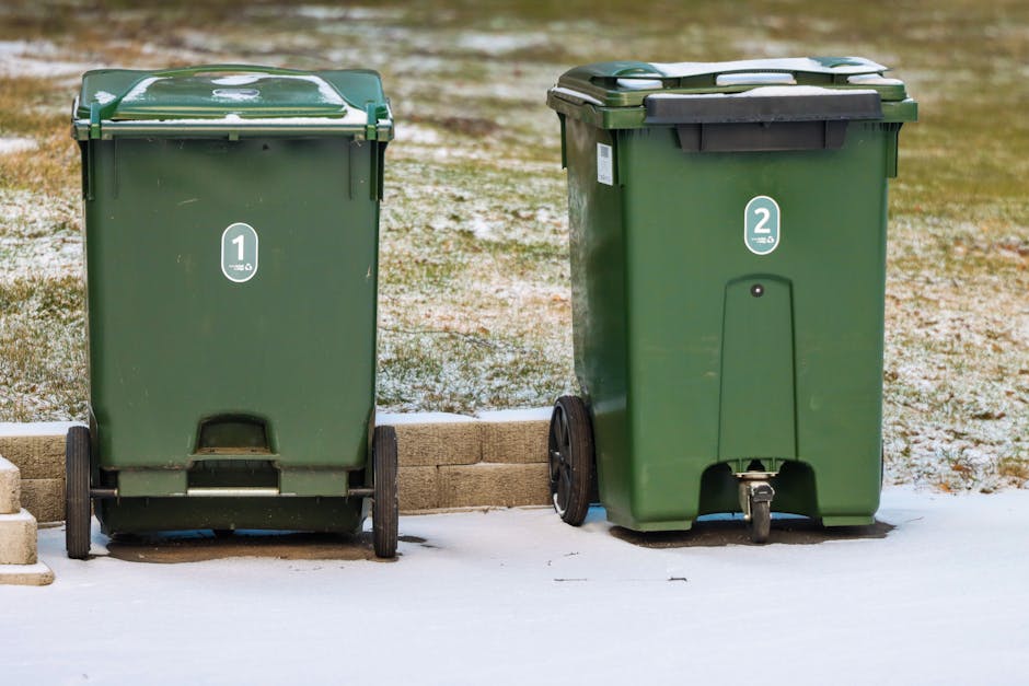 A black cylindrical rubbish bin made of metal with vertical ridges and a small lockable door on the front, positioned on a cobblestone street, is heavily surrounded by overflowing litter. The top of t
